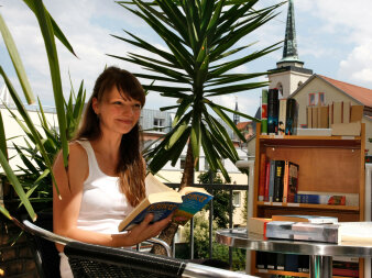 Eine Frau liest auf der Terrasse ein Buch. Im Hintergrund sind Palmengewächse und ein Kirchturm.