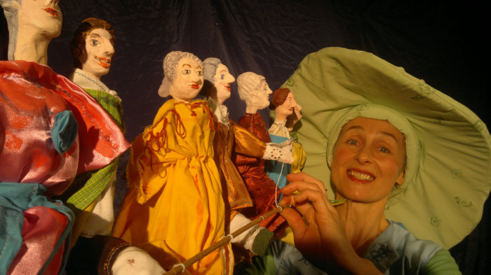 Theaterfirma in der Bibliothek Frau mit grünem Hut, vor ihr 6 Puppen.