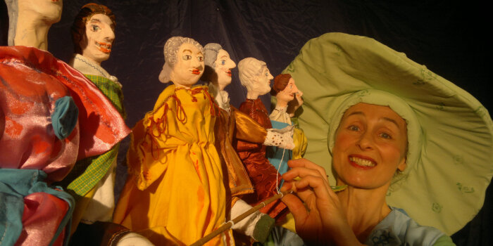 Frau mit grünem Hut, vor ihr 6 Puppen.