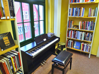 Zwischen zwei Bücherregalen und direkt vor dem Fenster steht ein schwarzes Piano.