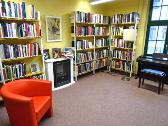Die Wände sind mit Bücherregalen versehen. Ein roter Sessel, ein Kamin und ein Piano schmücken den Raum.