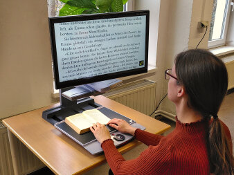 Eine Frau sitzt vor einem Bildschirm mit stark vergrößerter Schrift, darunter befindet sich ein aufgeklapptes Buch.