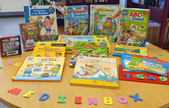 Ein Tisch voller diverser Medien für Vorschulkinder.Ausgeschnittene Buchstaben ergeben das Wort "Medienbox".