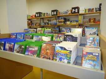 Im Vordergrund befinden sich zahlreiche Konsolen- und PC-Spiele. Im Hintergrund sind die Gesellschaftspiele-Regale.