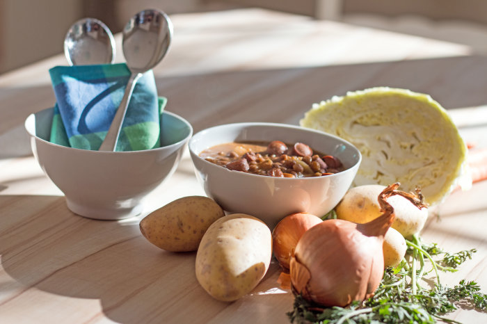 Suppen-Kochkurs Zwei kleine weiße Schüsseln auf hellem Küchentisch. Eine ist gefüllt mit heißer Linsensuppe. In der anderen Schüssel befinden sich, eingewickelt in eine Serviette, zwei Löffel. Im Vordergrund liegen auf dem Küchentisch: Möhren, Zwiebeln, Kartoffeln und ein Krautkopf.