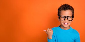 Vor orangem Hintergrund steht ein kleiner Junge mit blauem Pullover und zeigt mit dem Daumen nach rechts.