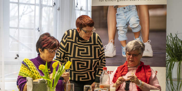 Zwei Damen stricken sitzend an einem Tisch, eine Dame unterhält sich mit den beiden.