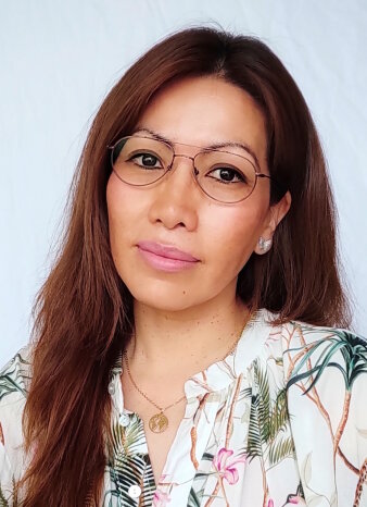 Eine Frau mit langen rotbraunen Haaren, einer feinen Brille und einer floralen Bluse blickt in die Kamera.