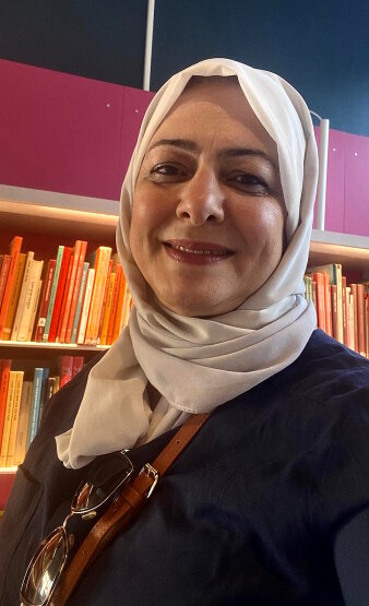 Eine Frau mit einem beige farbenen Kopftuch steht vor einem Bücherregal und lächelt in die Kamera