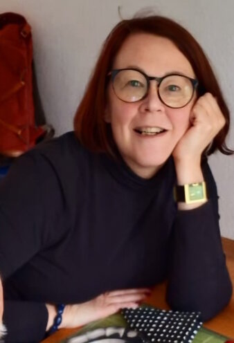 Eine Frau mit halblangen roten Haaren und Brille lächelt in die Kamera.