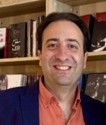 Ein Mann mit orangem Hemd und blauen Sakko steht vor einem Bücherregal und lächelt