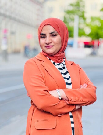 Eine Frau mit orangener Jacke und einem Kopftuch steht auf dem Erfurter Anger und lächelt in die Kamera