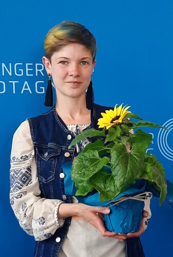 Eine junge Frau mit bestickter Bluse, blauer Weste und einer Sonnenblume in der Hand lächelt in die Kamera.