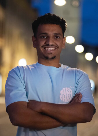 Ein Mann im hellblauen T-Shirt und verschränkten Armen lacht in die Kamera.