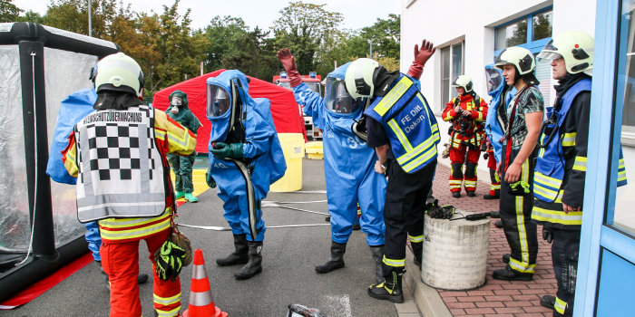 Feuerwehrleute rüsten sich mit blauen Schutzanzügen aus. Rundherum stehen Geräte für die Dekontamination.