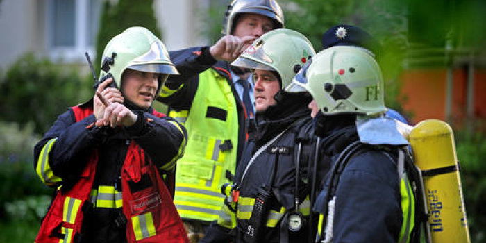 Das Foto zeigt einen Zugführer, der zwei Feuerwehrleute bei einem Feuerwehreinsatz zur Brandbekämpfung den Einsatzauftrag erläutert. Der Bildhintergrund zeigt den Einsatzleiter während einer Abstimmung mit Polizeikräften.