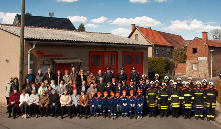 Angehörige der Feuerwehrwehr (Altersabteilung, Jugendfeuerwehr, Einsatzabteilung) vor dem Feuerwehrhaus (Flachbau mit 2 Toren)