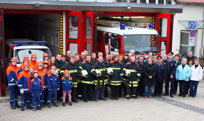 Feuerwehrangehörige (Jugendfeuerwehr, Einsatzabteilung, Altersabteilung) und zivile Vereinsmitglieder stehen vor dem Feuerwehrhaus mit 2 offenen Toren, in denen Feuerwehrfahrzeuge stehen