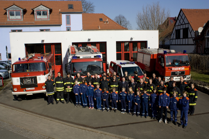 Kameradinnen und Kameraden der Feuerwehr vor dem Feuerwehrhaus mit 3 Toren und 4 Feuerwehrfahrzeugen