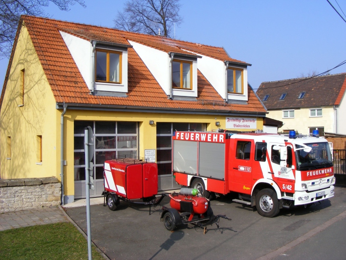 Feuerwehrhaus mit zwei Toren, davor Löschfahrzeug und zwei Anhänger