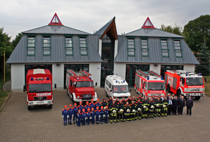 Feuerwehrangehörige vor dem Feuerwehrhaus mit 4 Toren und 5 Feuerwehrfahrzeugen