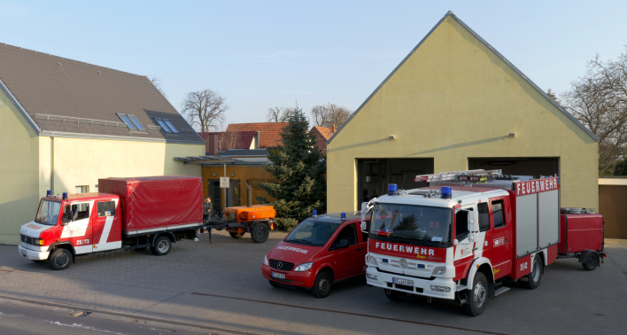 Drei Feuerwehrfahrzeuge und 2 Anhänger stehen vor einem gelben Feuerwehrhaus