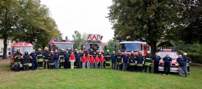 Feuerwehrangehörige in Einsatzbekleidung sowie Jugendfeuerwehrangehörige und Alterskameraden stehen vor 5 Feuerwehrfahrzeugen auf einer Wiese mit Bäumen