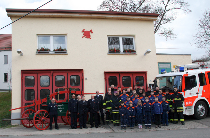 Feuerwehrangehörige der Altersabteilung, der Jugendfeuerwehr und der Einsatzabteilung stehen vor den Feuerwehrhaus mit zwei Toren und einem Obergeschoss, rechts daneben steht ein Feuerwehrfahrzeug