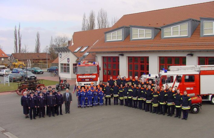 Feuerwehrangehörige der Alters- und Ehrenabteilung, Jugendfeuerwehr und Einsatzabteilung mit Feuerwehrfahrzeugen vor dem Feuerwehrhaus mit 4 Toren