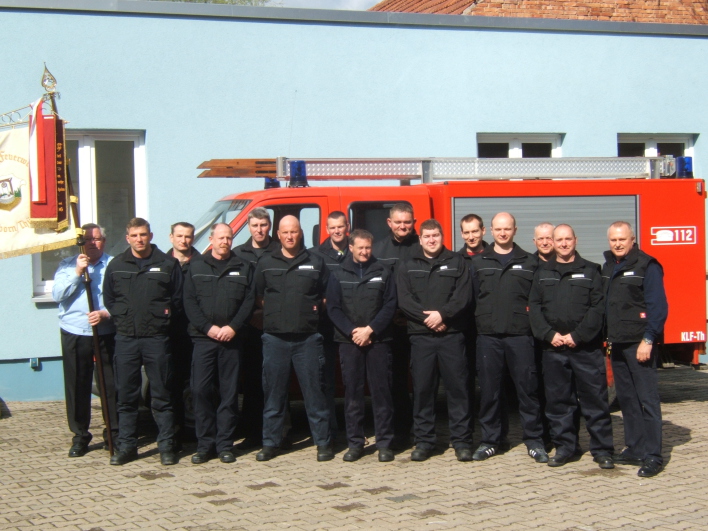 Feuerwehrangehörige stehen vor einem Feuerwehrfahrzeug (Transportergröße) vor einem Gebäude. Ein Feuerwehrmann hält eine Fahne.