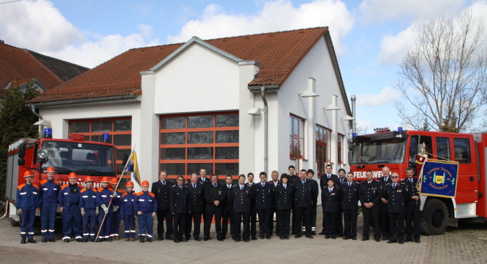 Feuerwehrangehörige in Uniform (Jugend, Einsatzabteilung und Altersabteilung) stehen mit 2 Fahnen vor zwei Feuerwehrfahrzeugen und dem Feuerwehrhaus mit 2 Toren