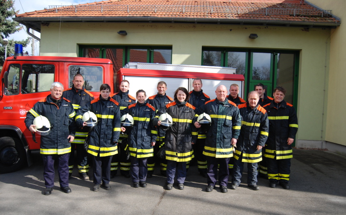 Feuerwehrangehörige in Einsatzbekleidung stehen vor einem Feuerwehrfahrzeug (Transportergröße) vor dem Feuerwehrhaus mit zwei Toren