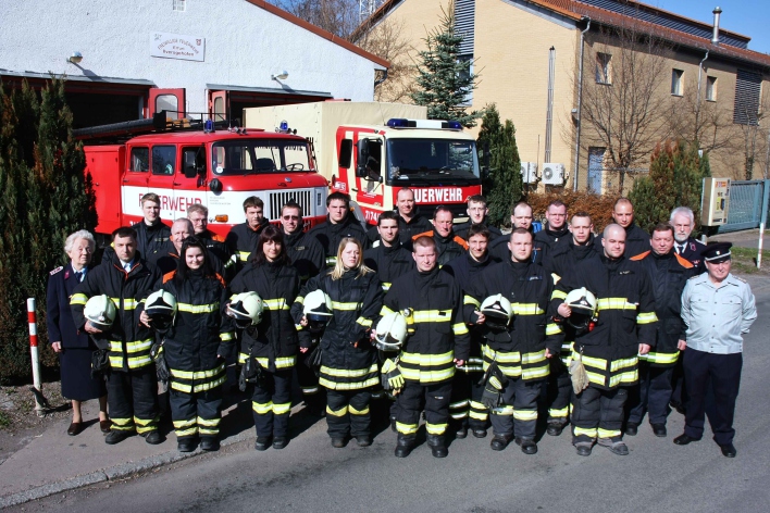 Feuerwehrangehörigen vor zwei Feuerwehrfahrzeugen und dem Feuerwehrhaus mit 2 Toren