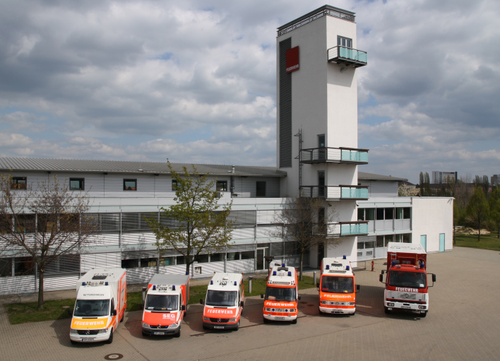 6 Feuerwehrfahrzeuge vor einem dreistöckigem Gebäude mit Schlauchturm