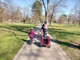 Ein Kind sowie eine Erwachsene fahren mit dem Rad durch den Park. An dem Rad der Frau ist ein Anhänger angebracht. 