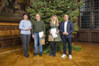 vier Personen stehen in einem festlich geschmückten Saal und halten Urkunden in der Hand
