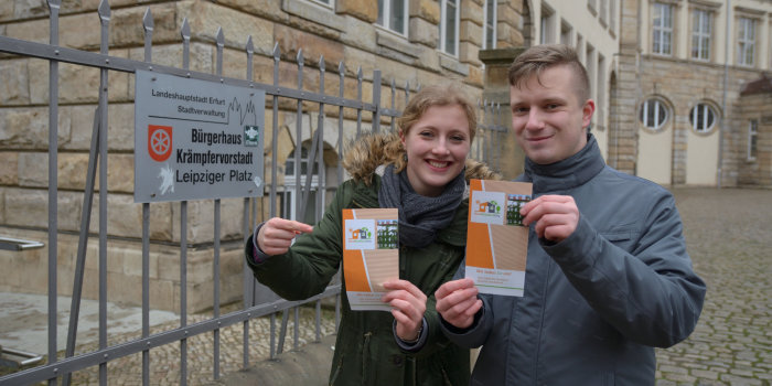 Junge Frau und junger Mann halten ein Faltblatt. Im Hintergrund ein großes Haus.