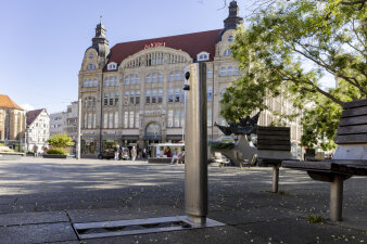 eine metallische Säule mit Wasserstrahl, im Hintergrund ein großes Gebäude