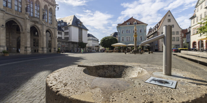 steinerner Trinkbrunnen auf einem Platz, im Hintergrund Altstadt-Kulisse