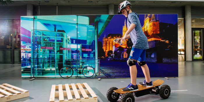 Foto: Leon Völker vom Staatlichen Gymnasium 10 in Erfurt © Steve Bauerschmidt Kind mit Helm auf Elekto-Skateboard vor Bildwand