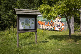auf einer Wiese stehen eine Infotafel aus Holz und ein Container mit Fuchs-Graffiti