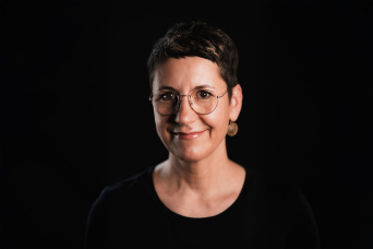 Eine Frau vor schwarzem Hintergrund schaut in die Kamera. Sie trägt eine Brille, hat kurze Haare und lächelt.