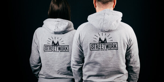 Foto: Streetwork Erfurt © Bert Billeb Ein Mann und eine Frau sind von hinten vor einem schwarzen Hintergrund zu sehen. Auf den Pullovern steht Streetwork