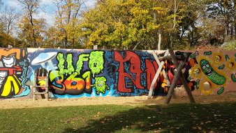 kleine Sandfläche mit Kleinkinderschaukel und großer bunter mit Graffiti bemalter Wand und Löchern zum Klettern und Durchgehen