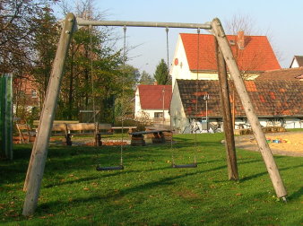 Rasenfläche mit großer Schaukel mit zwei Sitzen und große Sandfläche mit großem rot-gelb-grünen Kletterschloss aus Holz