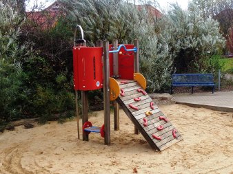 Sandfläche mit kleinem rot-gelb-blauen Kletterturm aus Holz mit Kletterrampe und Kletterstange