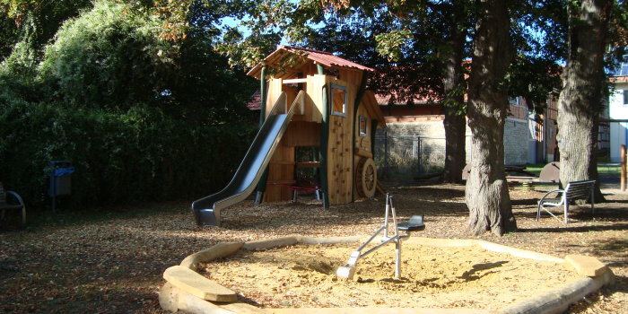 In Holz verkleidetes Turmhaus mit Rutsche. Im Vordergrund Sandkasten