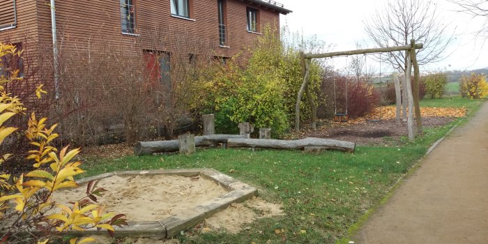 Rasenfläche mit kleinem Sandkaste, Balancierbalken aus Holz und einer Schaukel mit zwei Sitzen