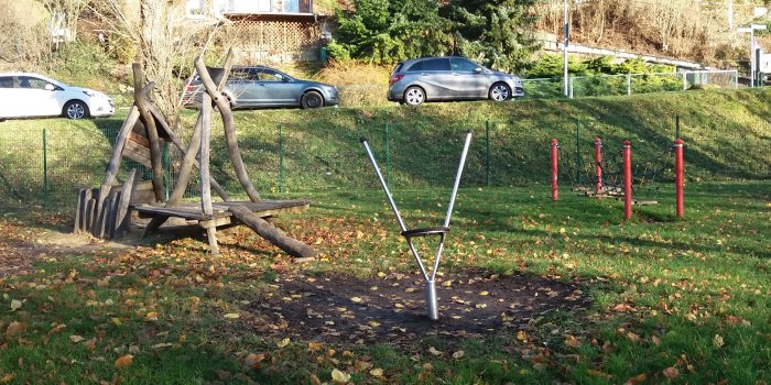 große Rasenfläche mit kleinem Drehtteller, roter Hängebrücke aus Holz und Seilen und Holzpfosten zum Sitzen und Klettern