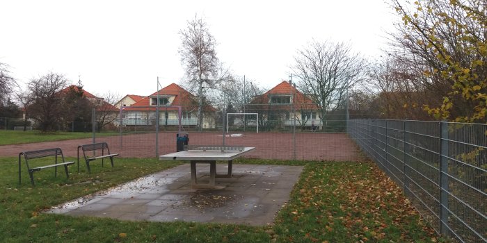 Tischtennisplatz mit Sitzbänken und eingezäunter Bolzplatz im Hintergrund